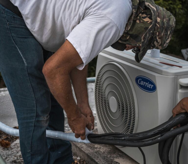 A technician performs maintenance on an outdoor air conditioning unit, focusing on hose connections.